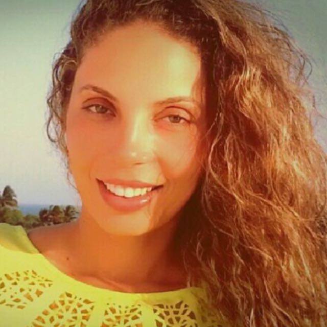 Uma mulher de cabelos cacheados está vestindo uma camisa amarela e sorrindo.