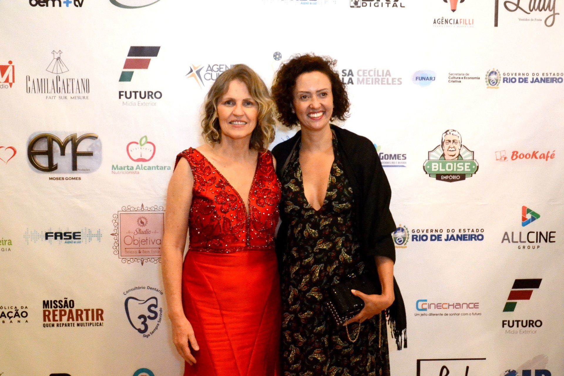 Duas mulheres estão posando para uma foto em frente a uma parede com logotipos.