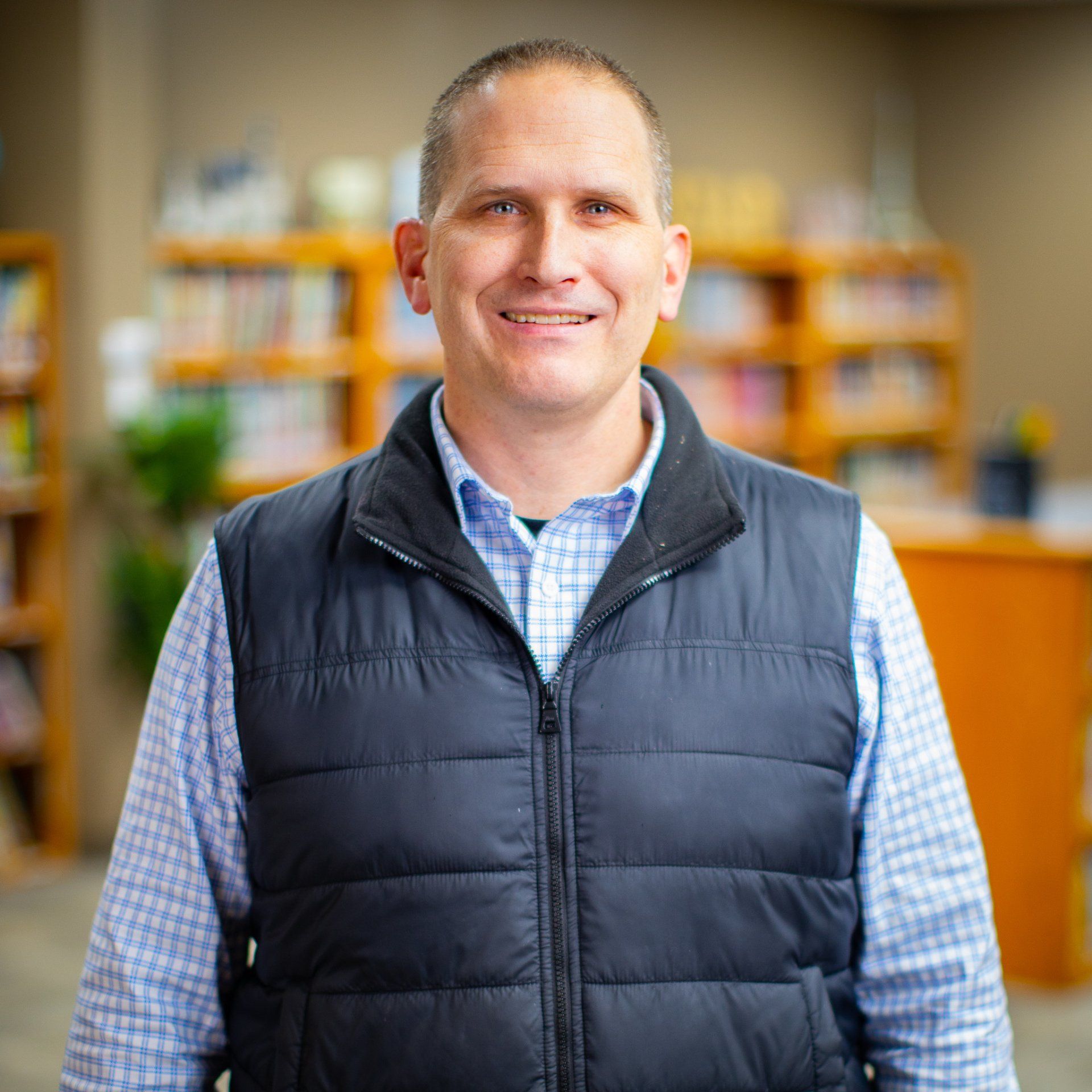 Smiling person in a navy vest and blue plaid shirt standing in a blurred indoor office or lobby setting