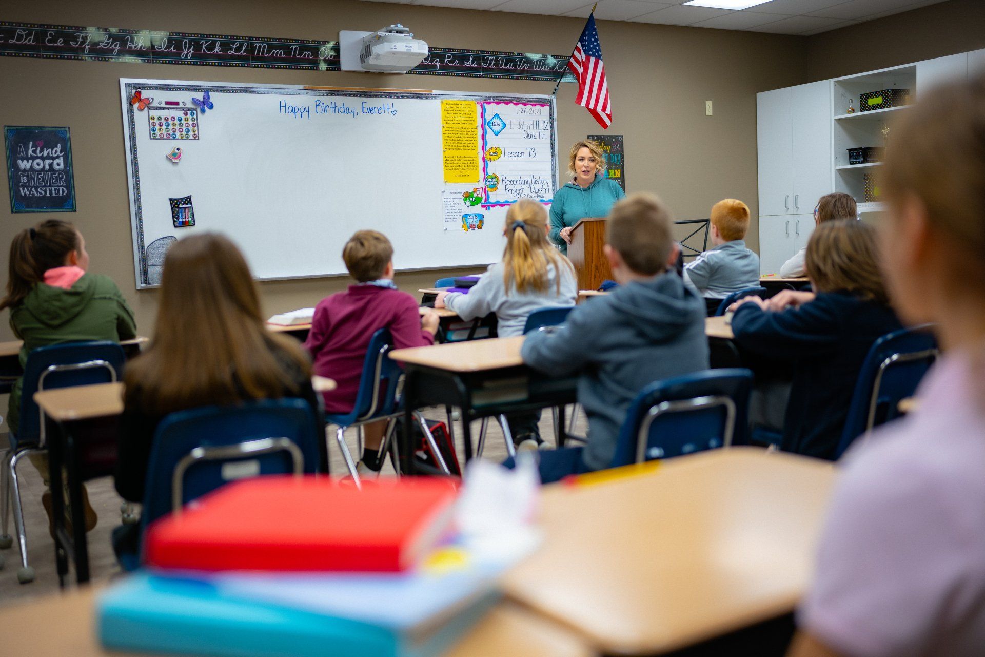 Classroom lesson with students seated at desks facing a teacher beside a whiteboard