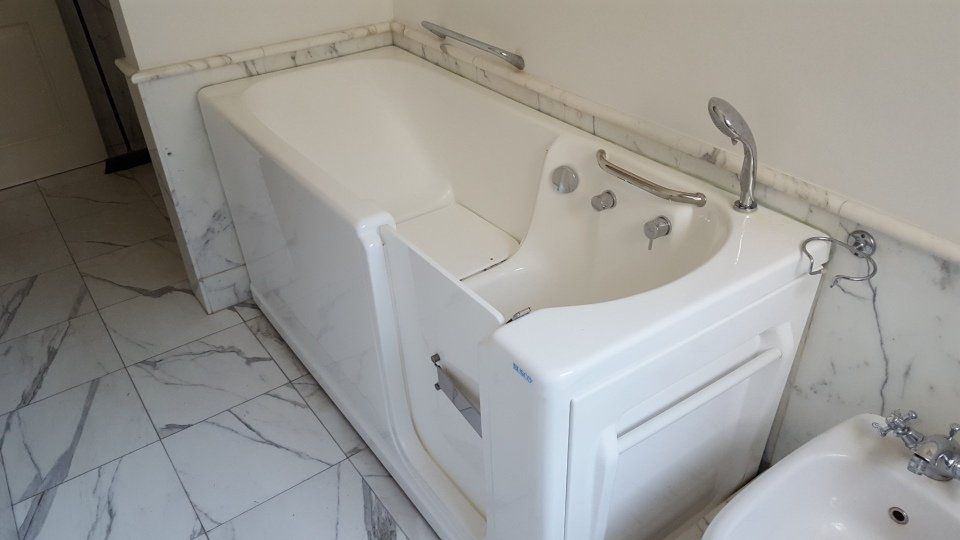 Vasca da bagno bianca walk-in con porta, inserita in un bagno con piastrelle e sanitari in marmo.