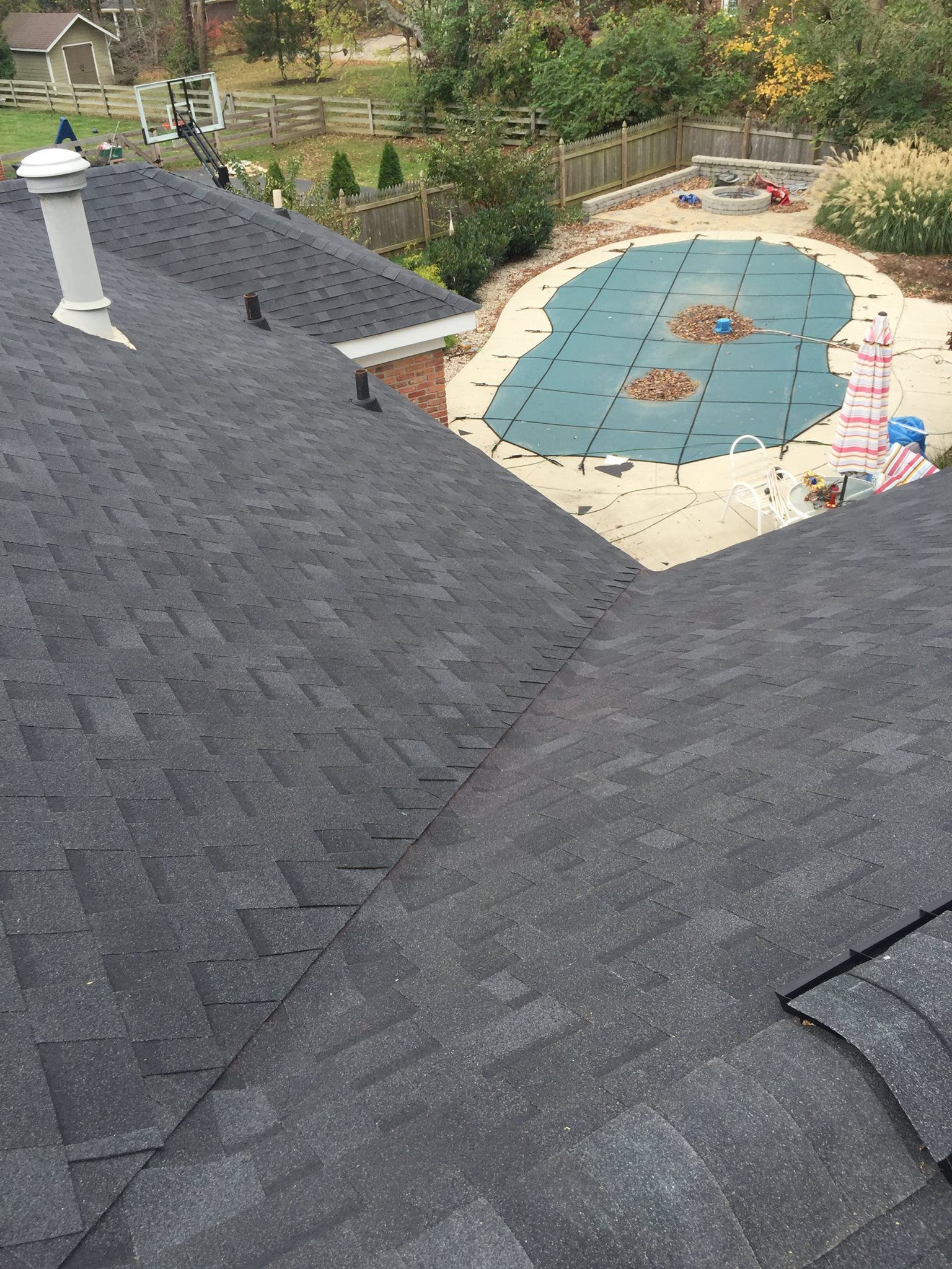 An elevated view of a gray asphalt shingle roof looking down toward a backyard swimming pool with a teal cover.