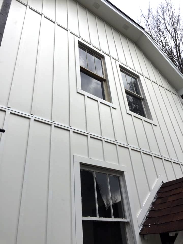 Low-angle view of a white, vertical-sided house exterior with three windows and a portion of a brown shingled roof.