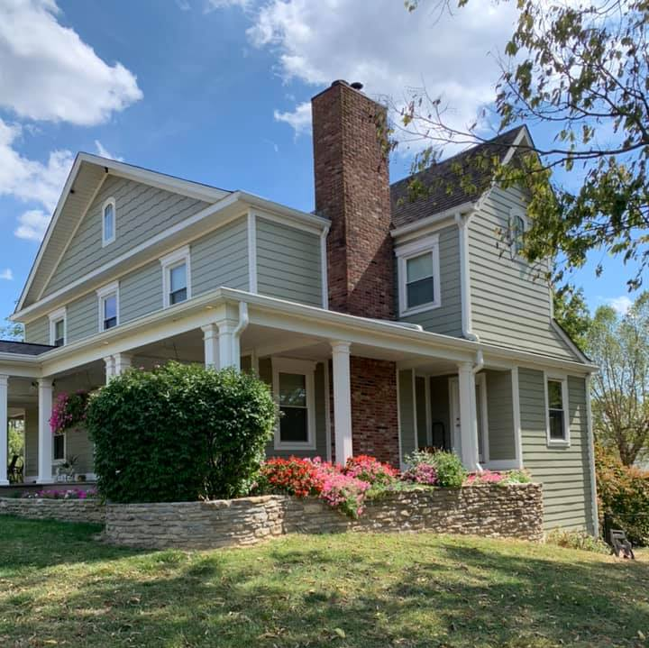 Two-story beige house with white trim, brick chimney, and a porch amid trees and flowering shrubs