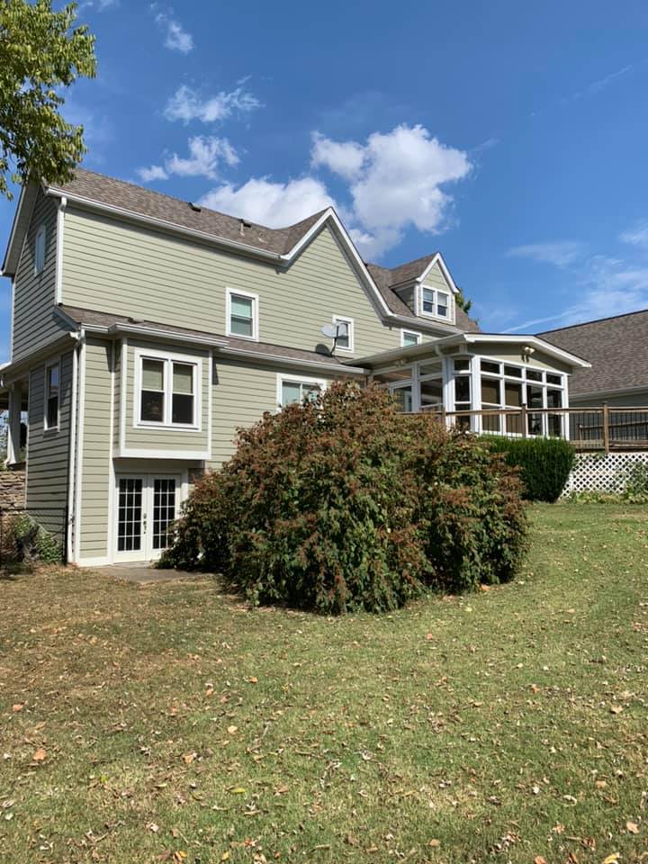 Two-story gray house with white trim, porch, and large bushes under a blue sky