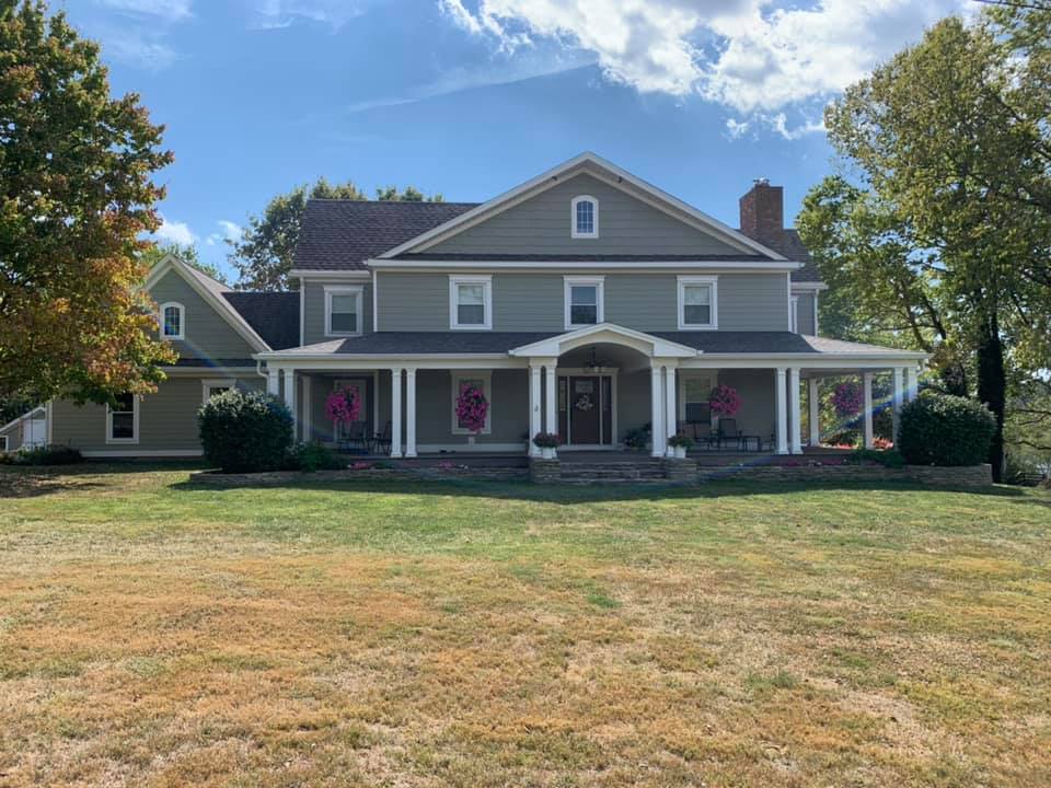 Large gray two-story house with white trim, a front porch, and red shutters on a sunny lawn.