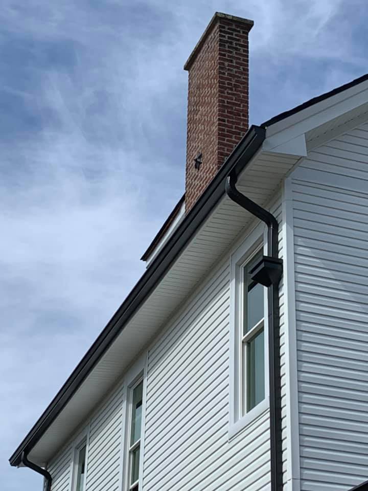 A low-angle view of a white-sided house with a brick chimney and black rain gutters against a blue, cloudy sky.