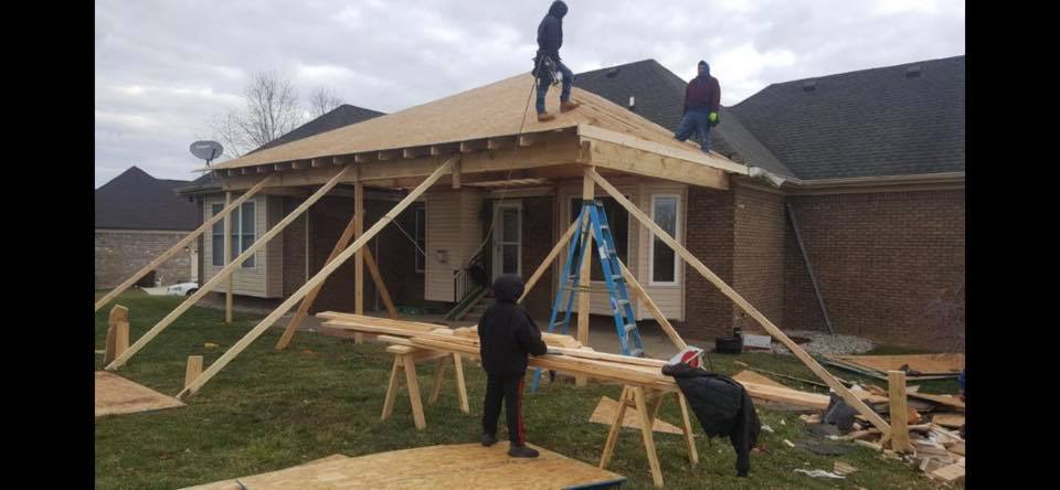 Workers framing a roof on a house, with wooden beams and ladders in a backyard.