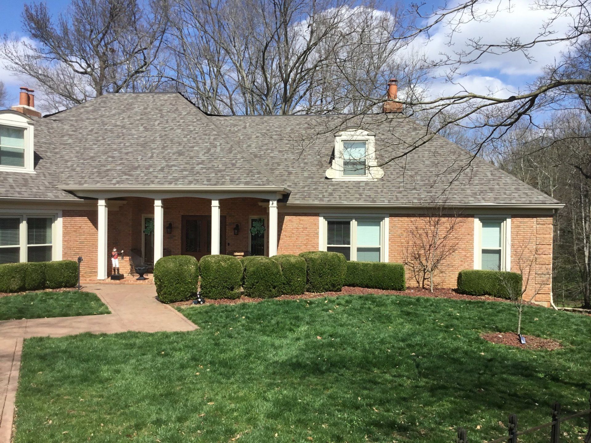 Brick suburban house with gray roof, white porch columns, green lawn, and bare trees under a blue sky