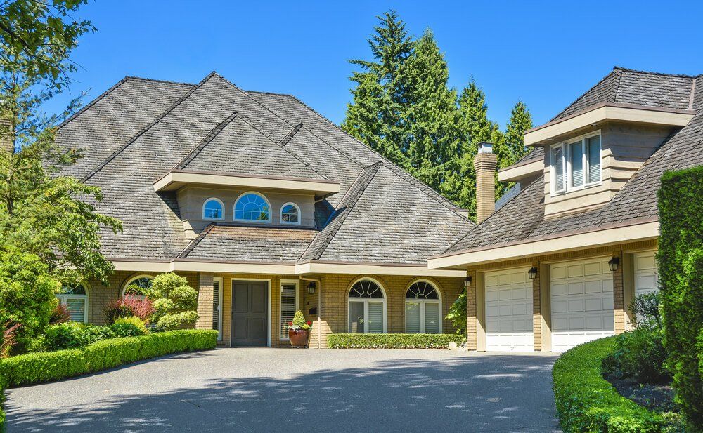 Large tan house with a gray shingled roof, a paved driveway, and manicured green hedges against a clear blue sky.