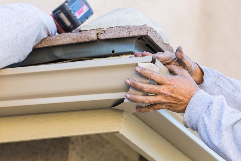 A worker in a light-colored long-sleeve shirt attaches a tan metal gutter end cap to the corner of a roofline.