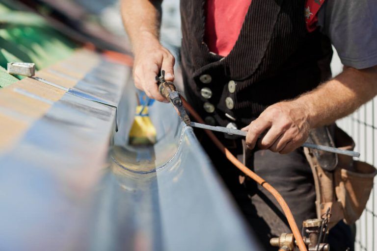 A tradesperson uses a soldering iron and solder to join metal gutters on a roof.