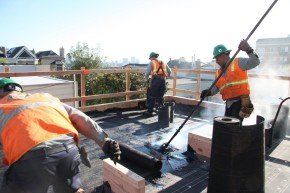 Roofers in safety vests applying hot tar to a flat roof on a sunny day.