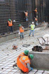 Construction workers in orange vests and hard hats install rebar on a concrete structure.