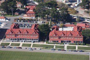 Red-roofed buildings with dormers in a grassy area, surrounded by trees and parked cars.