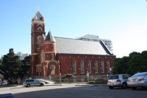 Red brick church with a tall steeple, on a sunny day. Cars are parked in the foreground.