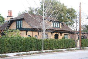 Cottage-style house behind a tall green hedge; brown roof, brick and dark trim.