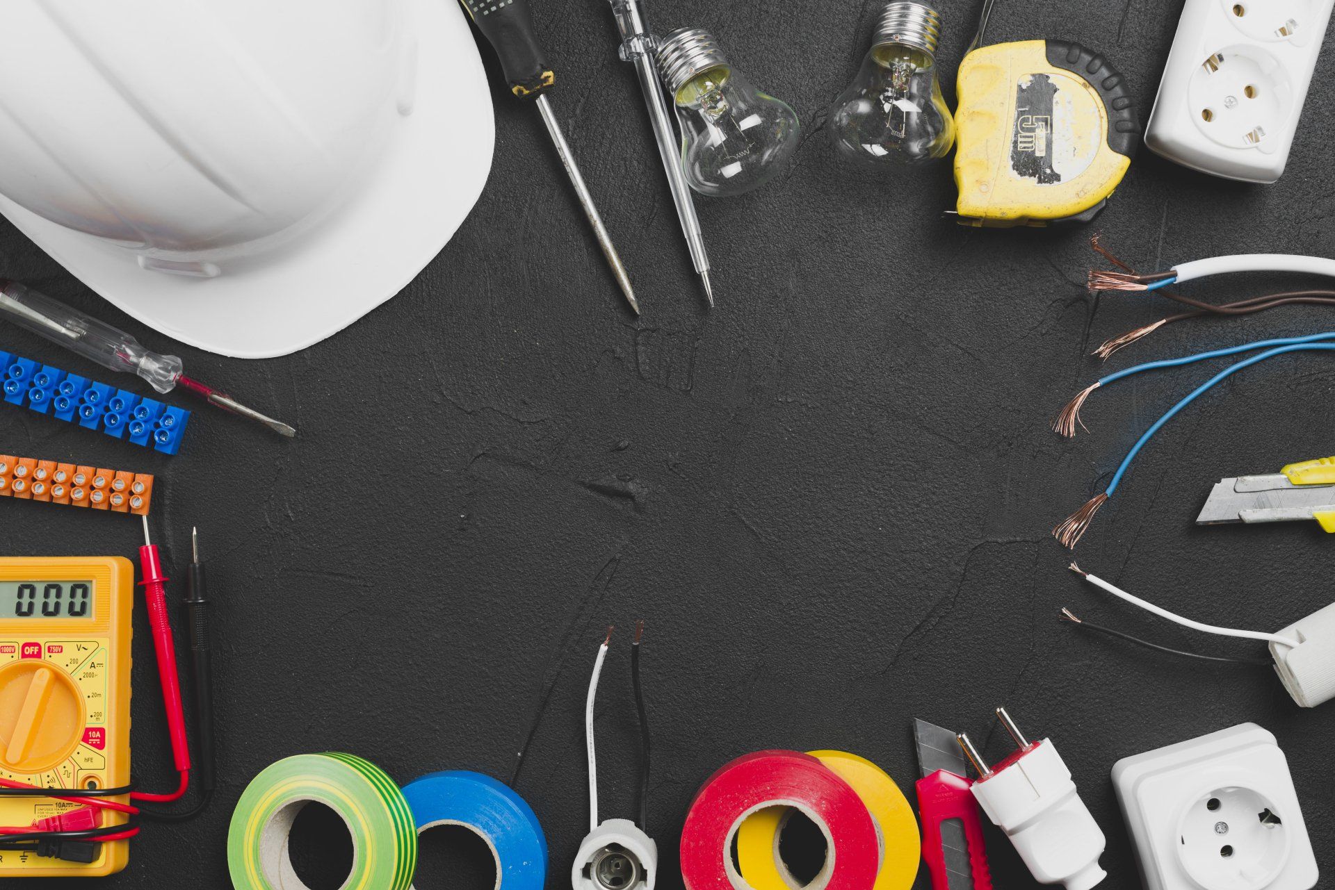 Electrician tools, wires, tape, outlets, and a hard hat arranged on a dark work surface.