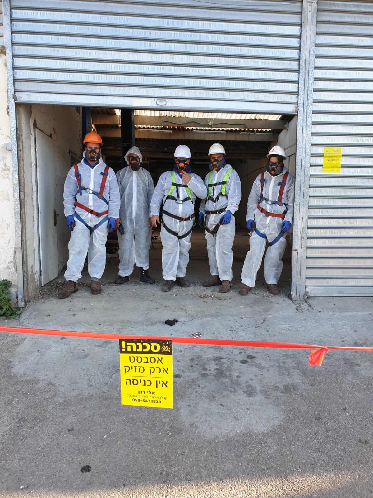 Five asbestos workers in safety gear stand at a warehouse doorway behind caution tape and a warning sign.