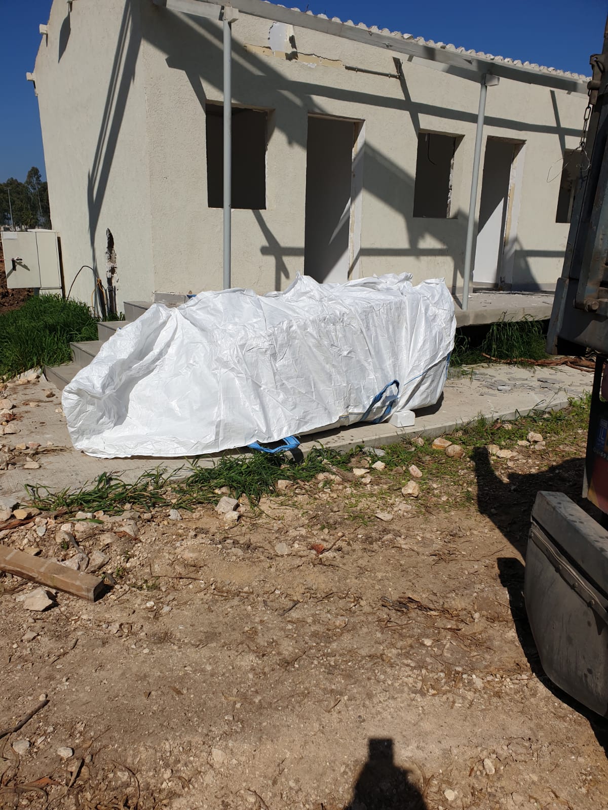 White tarp-covered object in front of a beige house with a porch and dry dirt yard.