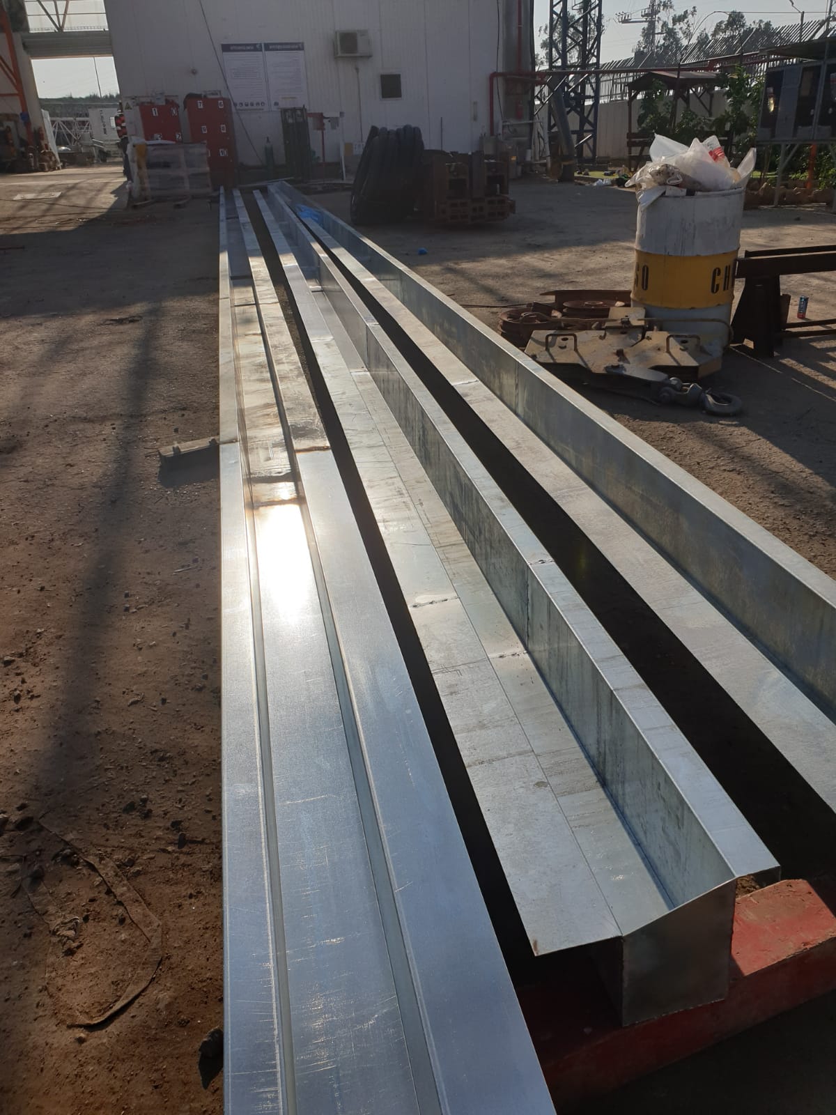 Stacked metal beams in an outdoor industrial yard, with sun reflecting off the long silver surfaces.