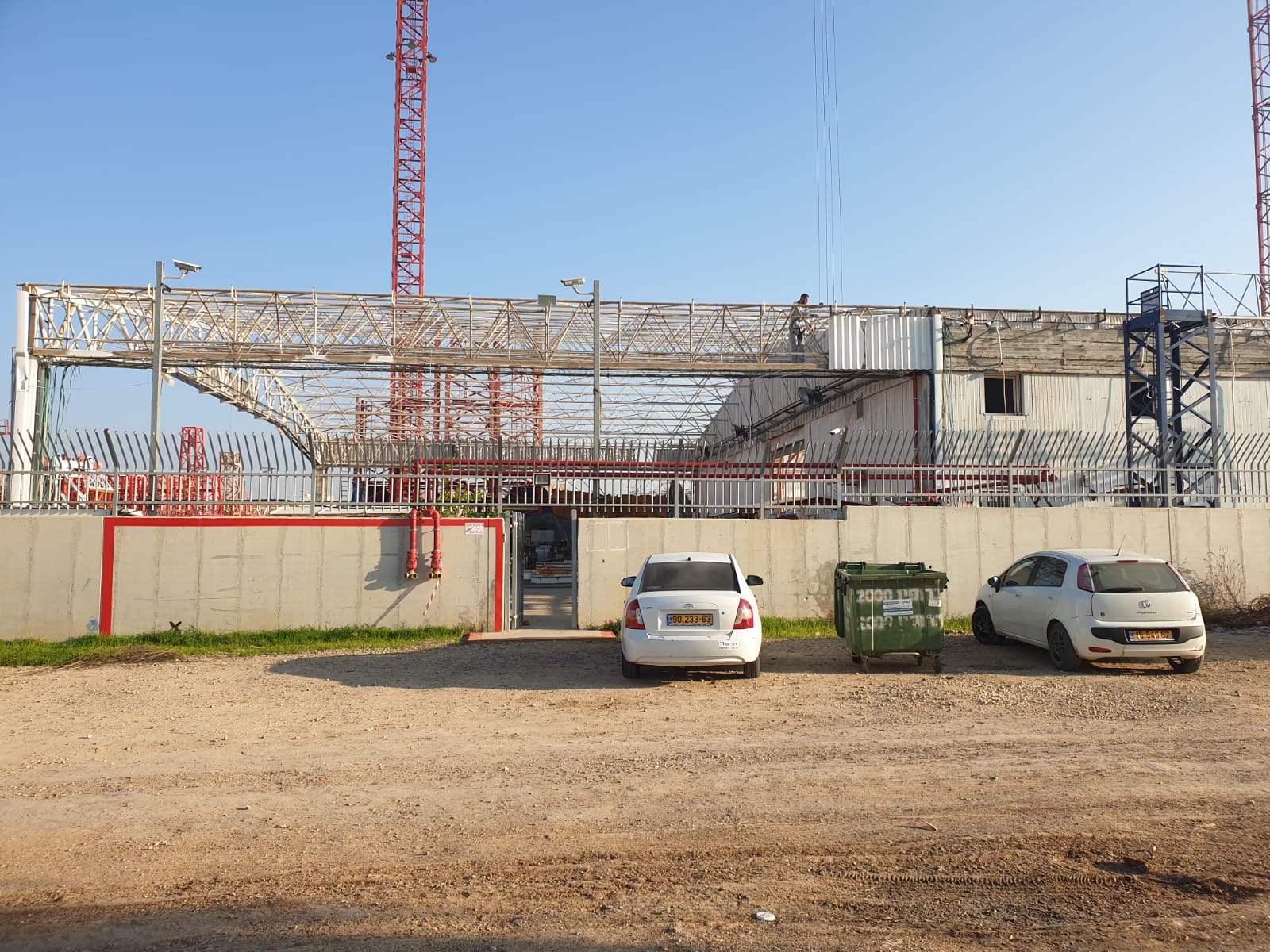 Construction site with steel framework, crane, and two parked cars behind a red gate.