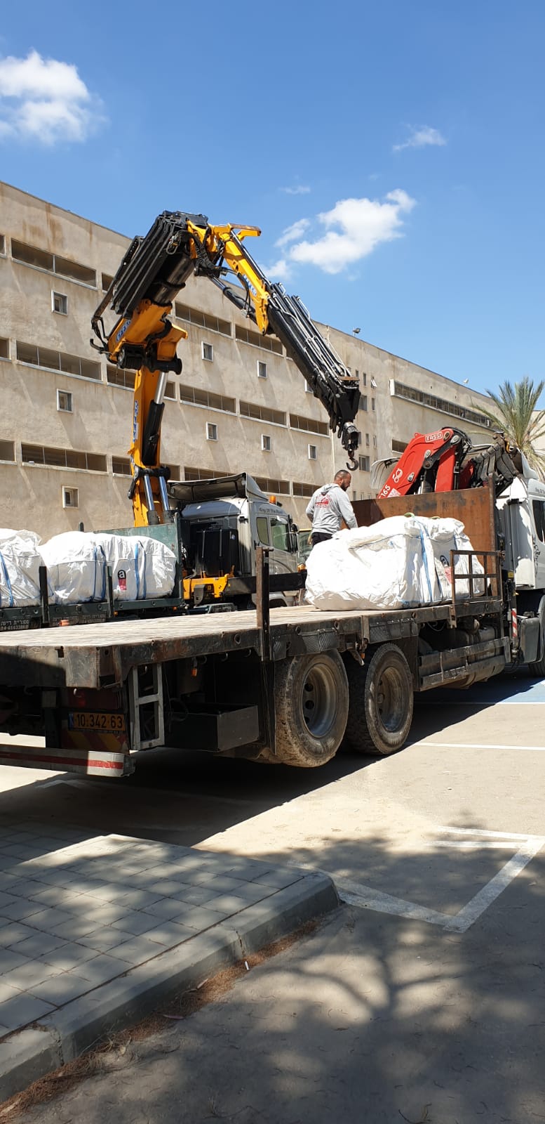 Flatbed truck loaded with white-wrapped cargo, with a crane arm lifting materials on a sunny street.