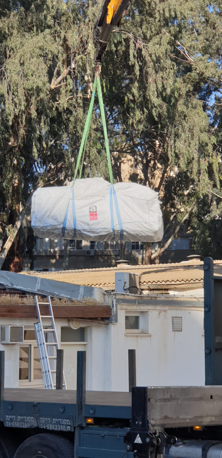 Crane hoisting a wrapped load above a building roof with green straps and trees in the background