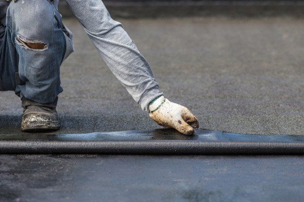Worker kneeling on pavement, smoothing a rolled strip of black roofing material with a gloved hand.
