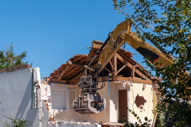 Excavator demolishing a house, witha sbestos  roof partially collapsed under a blue sky.