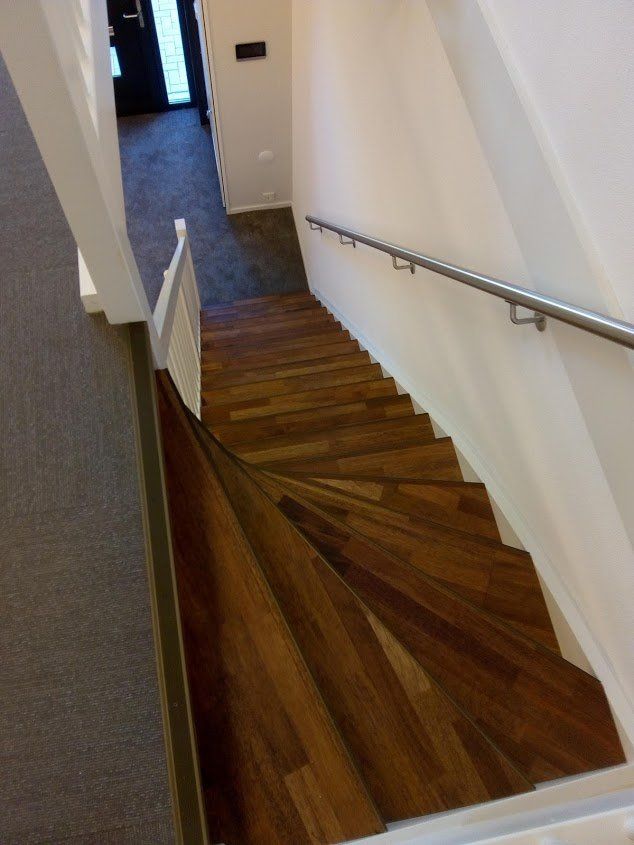 Een houten trap leidt vanaf een overloop naar beneden, met een metalen leuning tegen een witte muur en een witte balustrade aan de linkerkant.