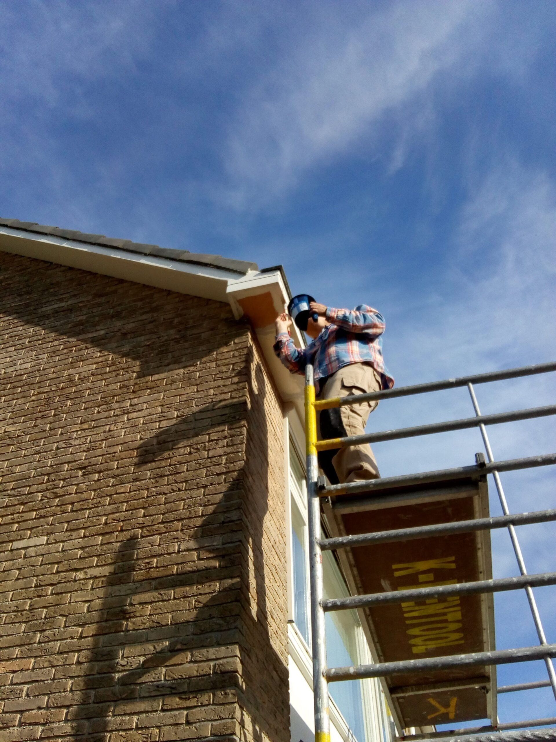 Een persoon in een geruit overhemd staat op een steiger en werkt aan de witte kozijnen van een bakstenen huis onder een blauwe hemel.