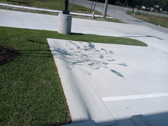 Sidewalk and parking lot corner with tree shadows, grass border, and a concrete post near a road.