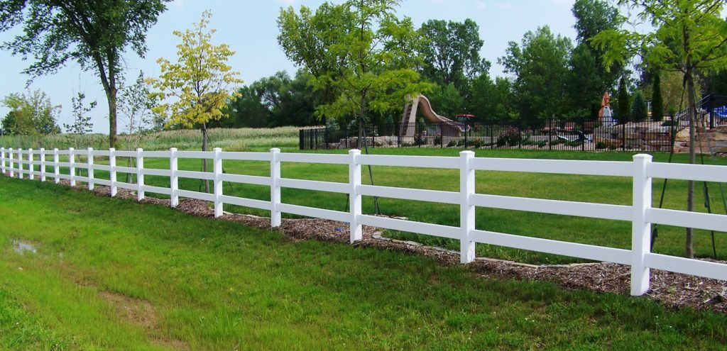 A split rail white vinyl fence surrounds a grassy field with trees in the background.