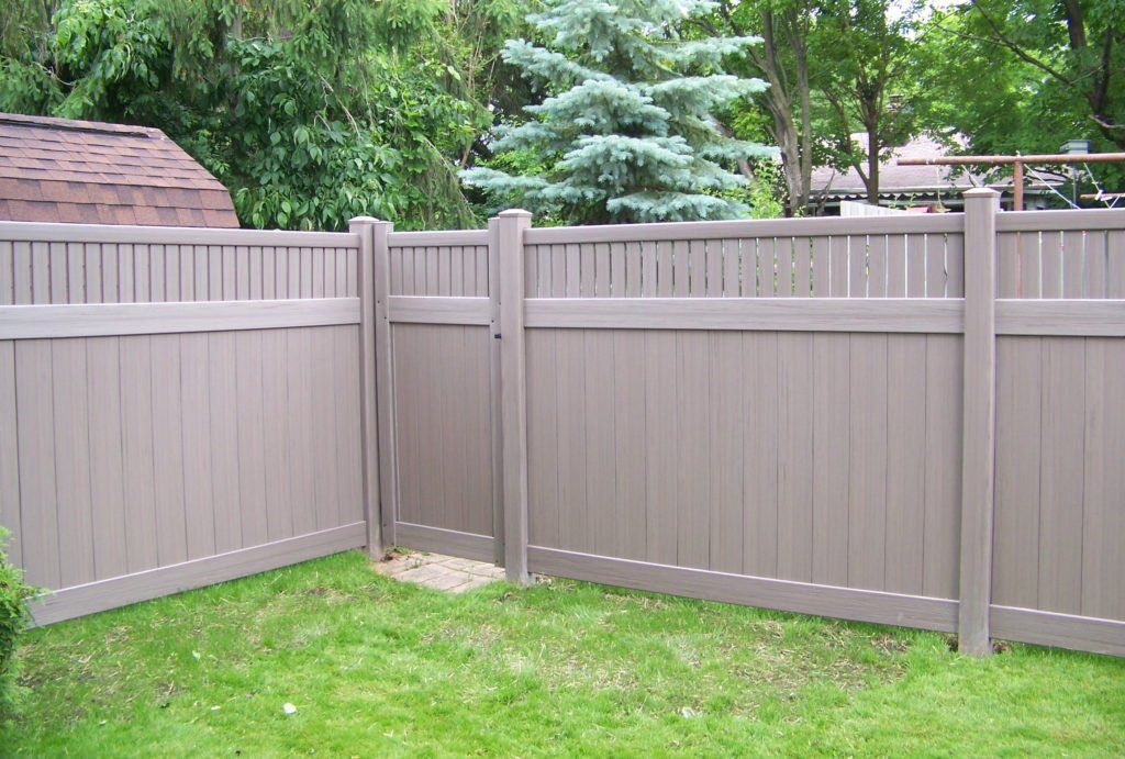 A brown vinyl fence is in the middle of a lush green yard with a gate