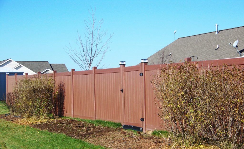 A vinyl fence is surrounded by bushes and a house in the background.