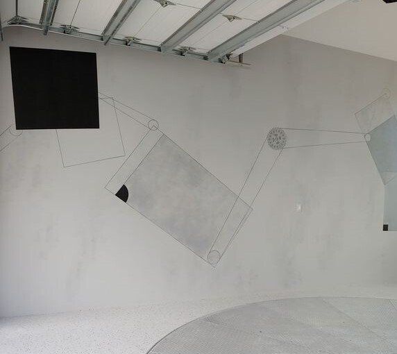 Gray room with a painted mechanical arm on the wall, a black square screen, and a partial circle on the floor.
