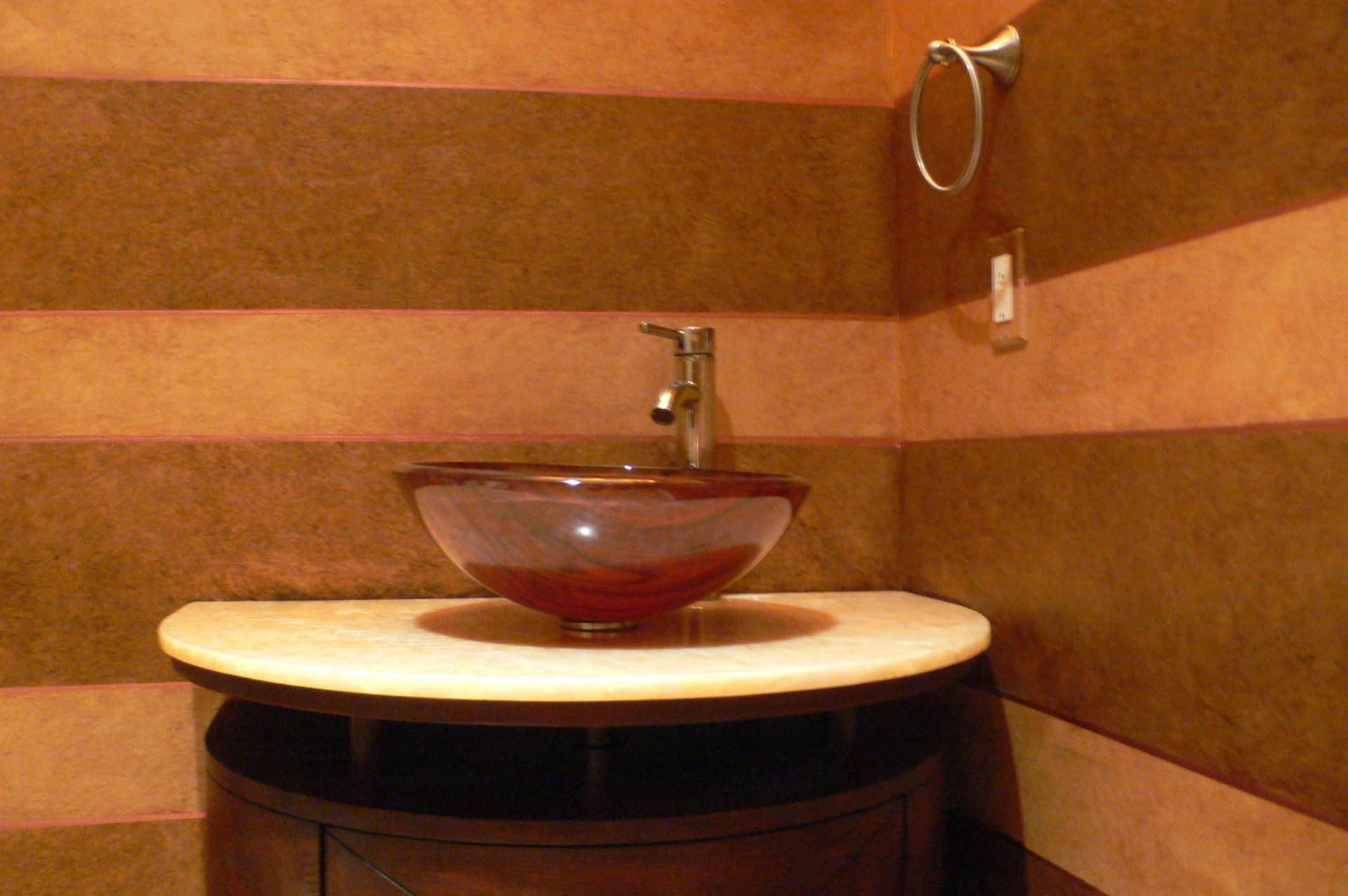 Brown vessel sink on a marble-like countertop, against striped brown walls, in a bathroom.