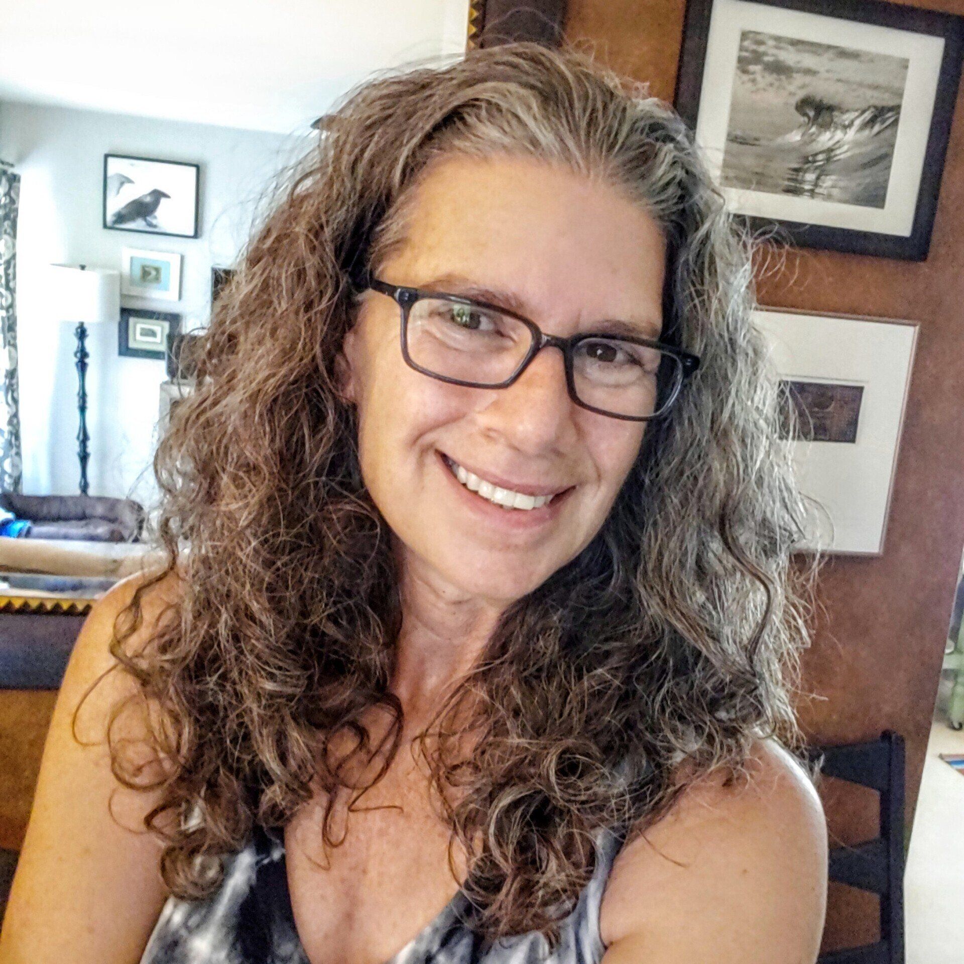 Woman with gray curly hair and glasses smiles, indoors, surrounded by art.