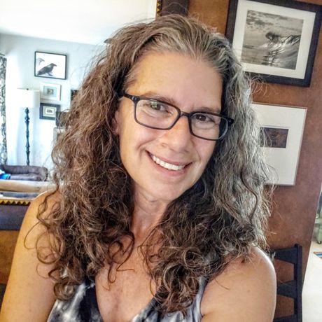 Woman with gray curly hair smiles, wearing glasses and a patterned top. Inside a room with artwork.