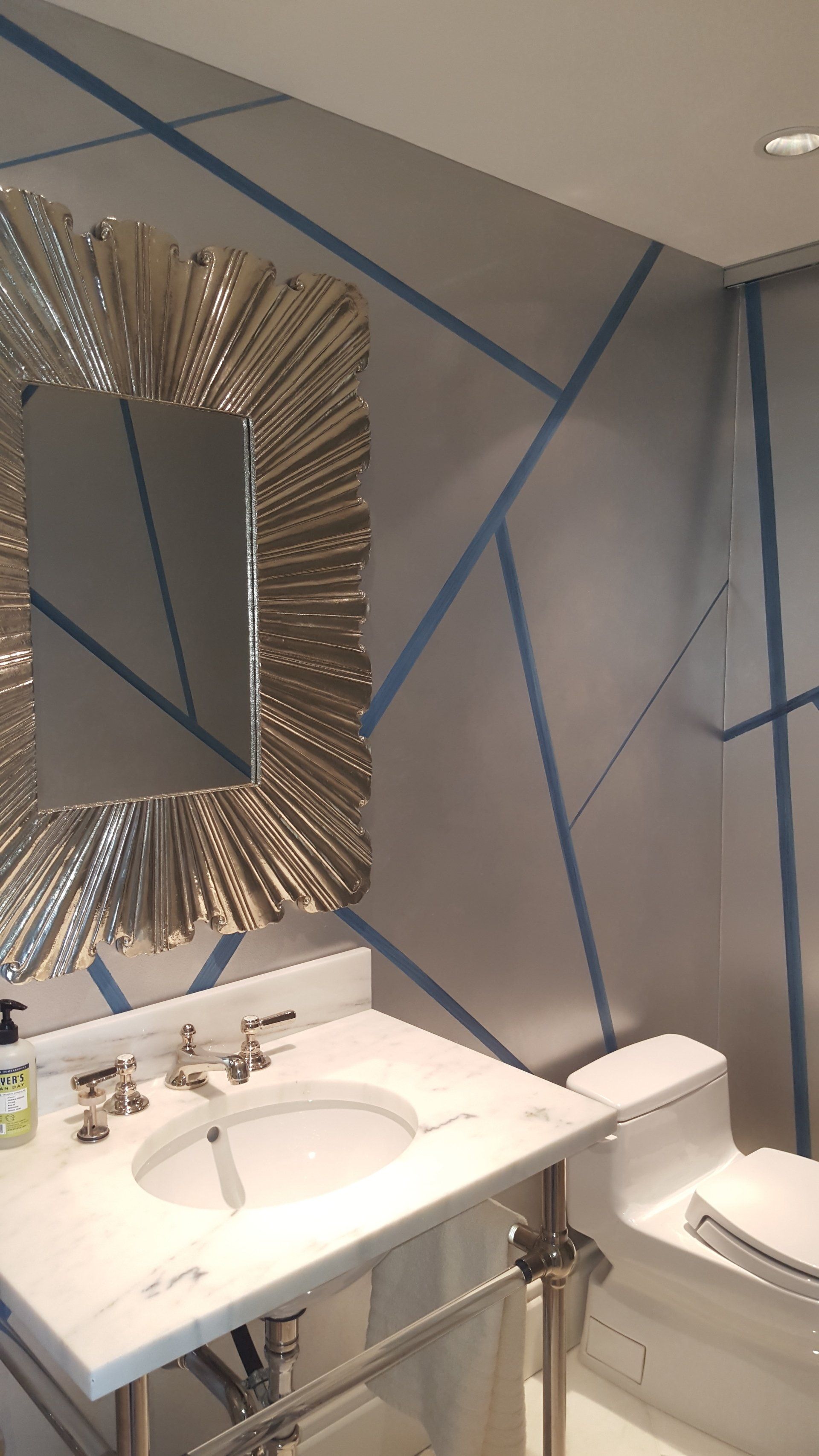 Bathroom with silver geometric wall design, ornate mirror, white sink, and toilet. Blue accent lines.