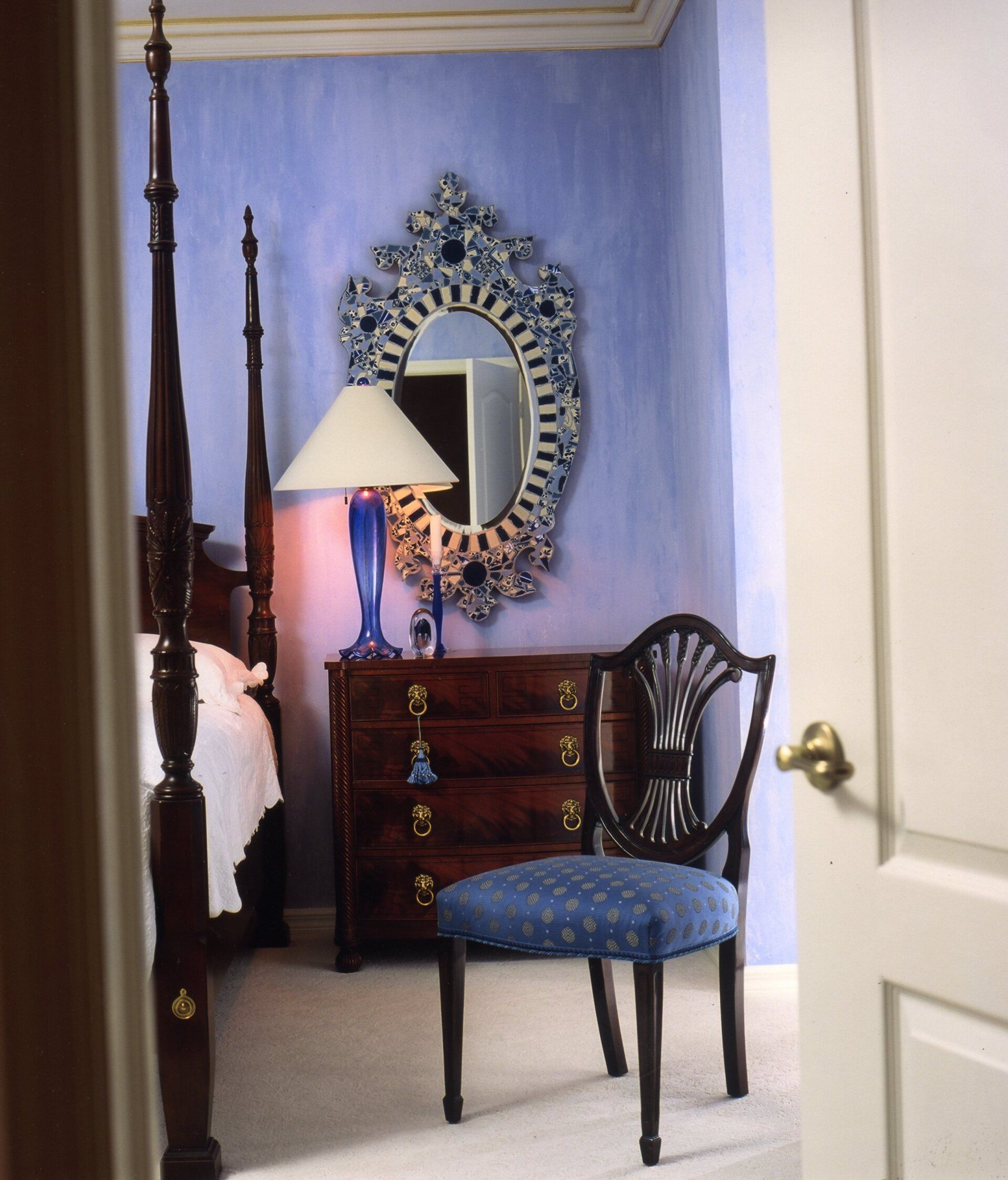 Bedroom interior: blue walls, ornate mirror, mahogany furniture, blue chair, white bedding, open door.