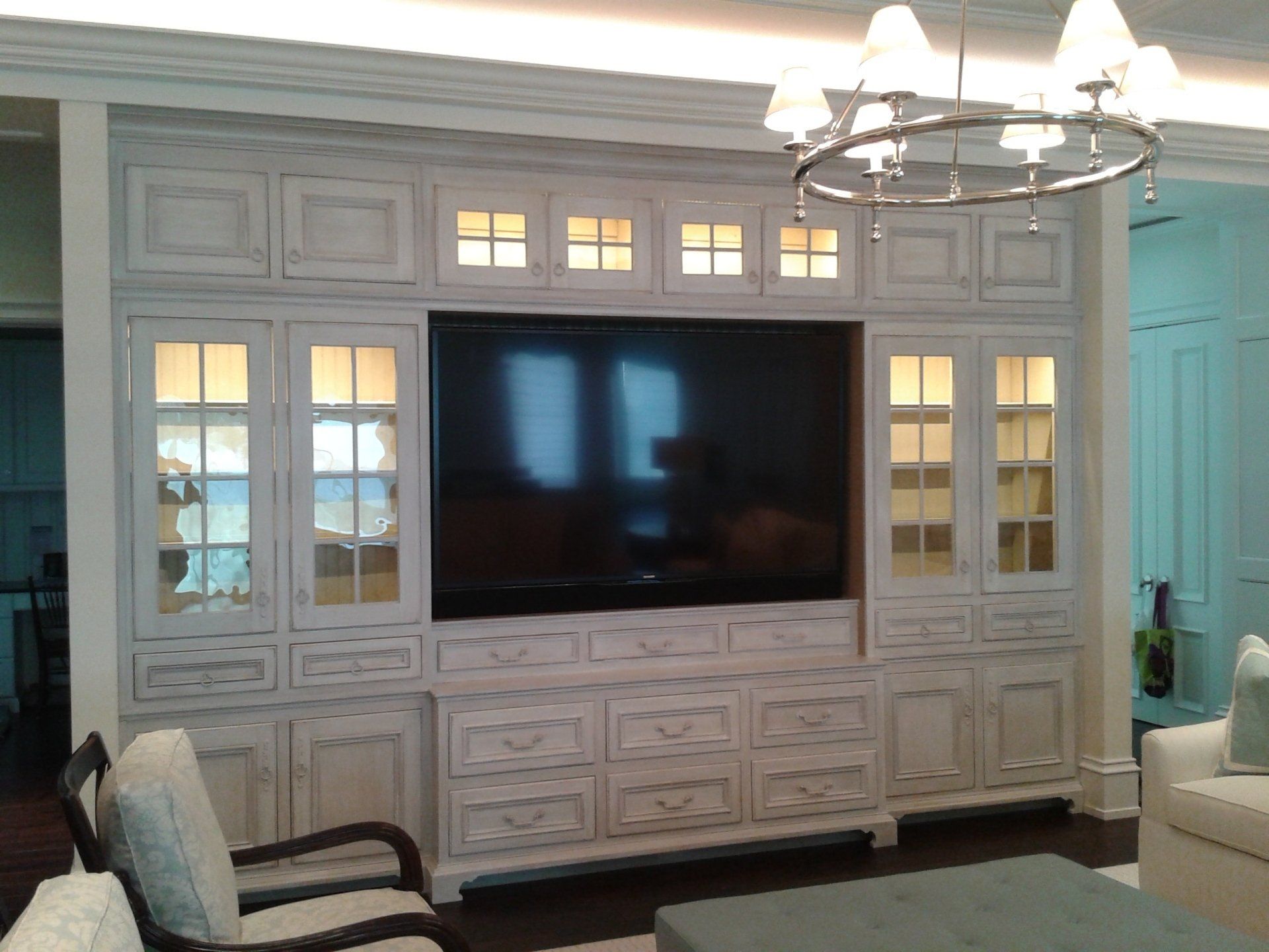 Built-in white cabinet with drawers, glass-door cabinets, and TV. Chandelier hangs nearby.