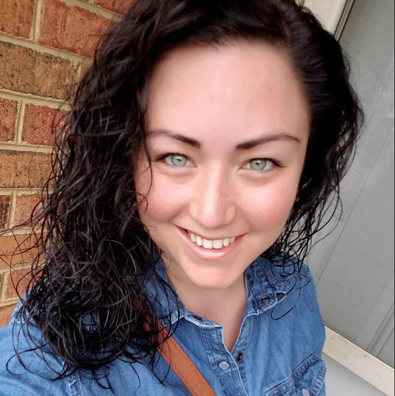 A woman in a denim shirt is smiling in front of a brick wall