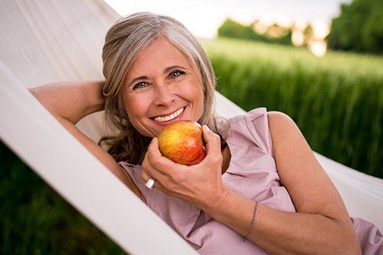 Woman with gray hair smiling, holding an apple, relaxing in a hammock outdoors.