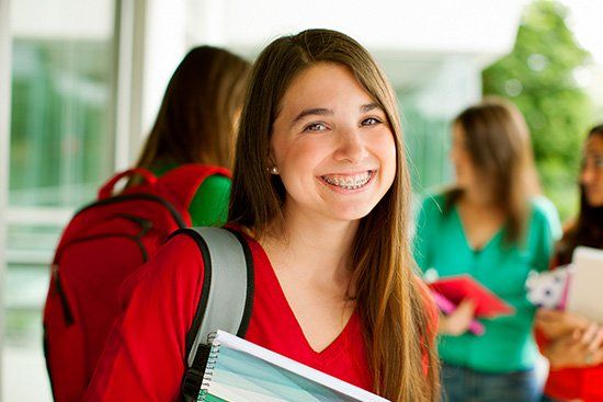 Teenage girl with braces smiling, wearing red shirt, backpack, holding notebooks, with blurred students in background.