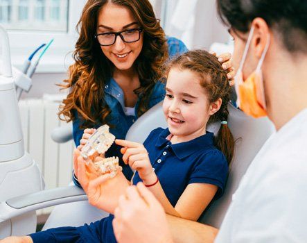 A dentist showing a young girl a model of teeth while her mother watches in the dental chair.