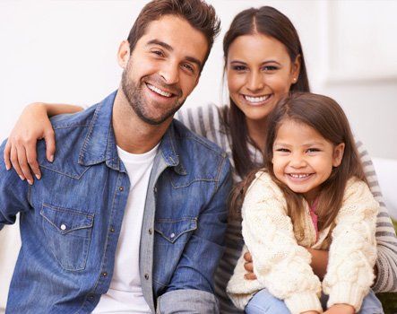 Family of three smiling at the camera. Father with arm around mother and daughter in their home.