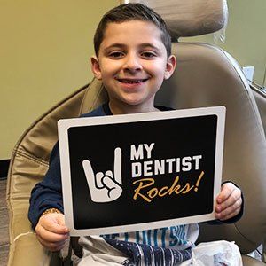 Young boy in dentist chair, smiling, holding sign: 