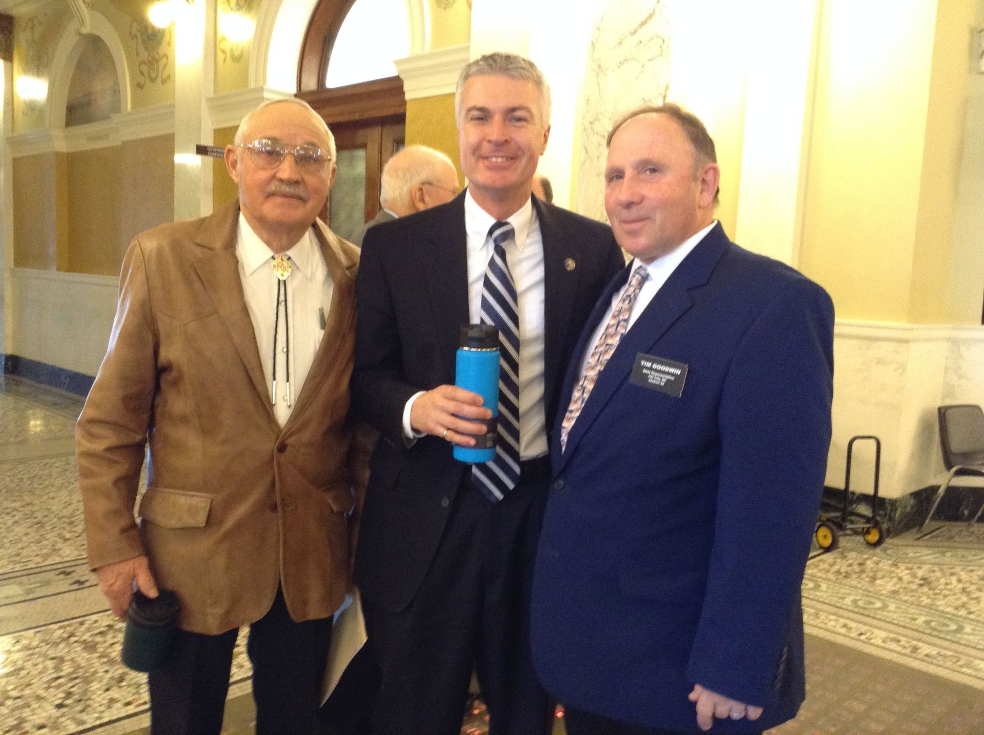 Three men standing together in a building with a water bottle; one wears a blue suit, one a tan jacket, and one a dark suit.